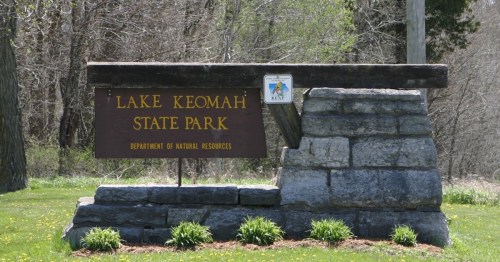 Sign for Lake Keomah State Park, featuring stone base and wooden top, surrounded by grass and trees.