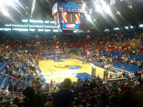 A wide view of a basketball arena filled with fans, featuring a court with the Kansas Jayhawks logo and scoreboard.