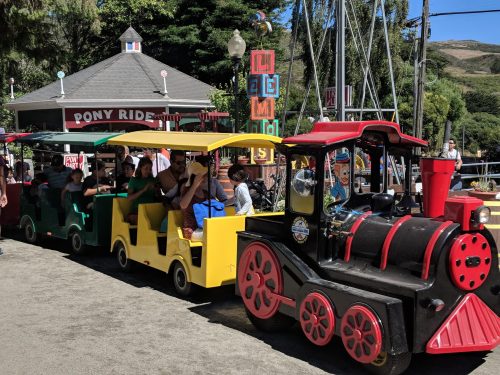 A colorful miniature train with passengers, parked near a pony ride attraction in a sunny outdoor setting.