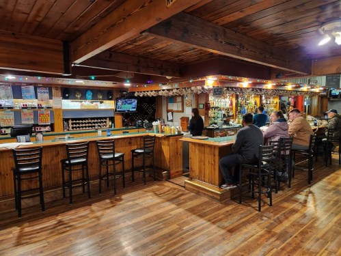 A cozy bar interior with wooden decor, patrons seated at the counter, and a TV displaying a sports game.