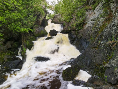 A cascading waterfall flows over dark rocks, surrounded by lush green foliage and trees.
