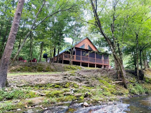 A cozy cabin nestled among trees by a stream, featuring a porch and steps leading down to the water.