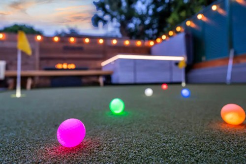 Colorful glowing golf balls on a putting green, with string lights and a sunset in the background.