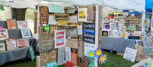 A vibrant outdoor market display featuring various handmade signs and crafts, with colorful decorations and tents.