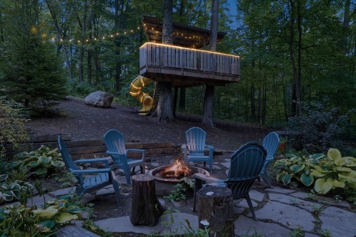 A cozy fire pit surrounded by chairs, with a treehouse above, illuminated by string lights in a serene forest setting.