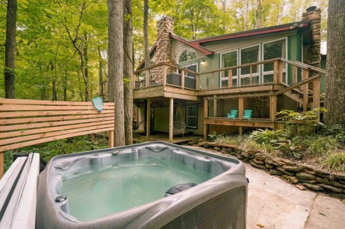 A hot tub in the foreground with a wooden deck and a house surrounded by trees in a serene forest setting.