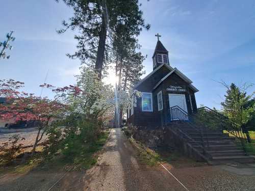 A small church with a steeple, surrounded by trees and blooming flowers, under a bright sun.