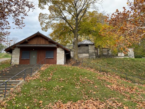 A rustic building with a wooden roof, surrounded by autumn leaves and trees, set on a grassy hillside.