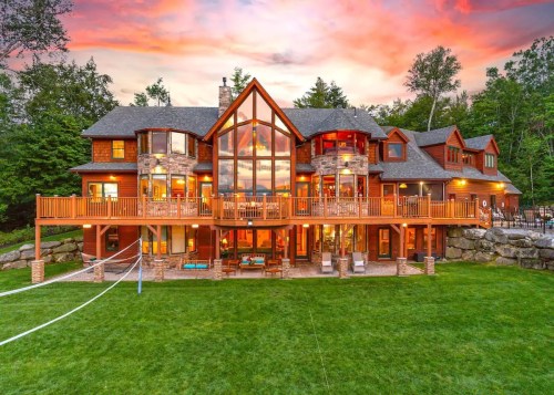 A large, modern house with a glass facade, surrounded by trees, at sunset, featuring a spacious deck and lawn.
