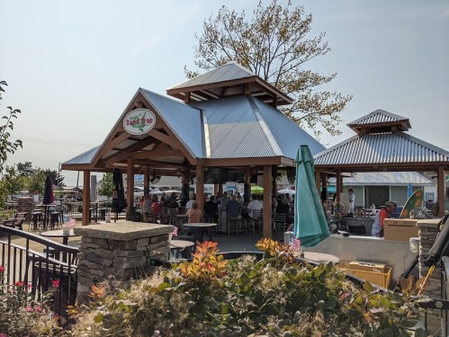 A wooden pavilion with a metal roof, surrounded by greenery and outdoor seating, bustling with people enjoying a sunny day.