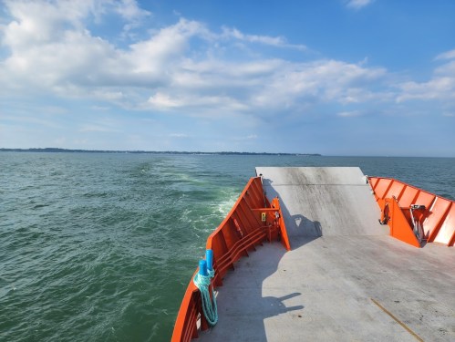 View from the bow of an orange boat, with calm waters and a distant shoreline under a partly cloudy sky.