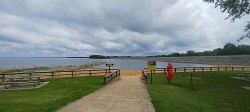 A sandy beach area by a lake, with picnic tables and a cloudy sky in the background. Safety signs are visible.