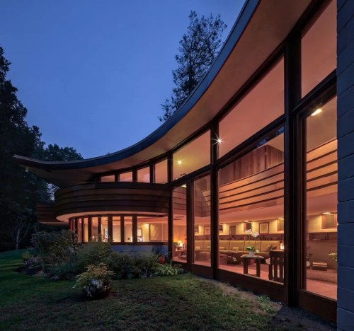 A modern curved house at dusk, featuring large windows that illuminate the interior with warm light.