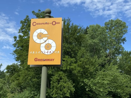 Sign for the Chesapeake and Ohio Greenway, featuring a colorful design and surrounded by green trees under a blue sky.