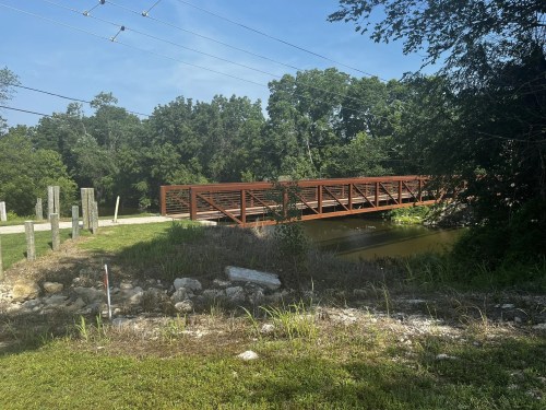 A red metal bridge spans a small body of water, surrounded by greenery and trees under a clear blue sky.