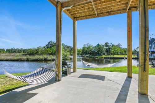 A serene outdoor space with two hammocks by a calm waterway, shaded by a wooden structure and surrounded by greenery.