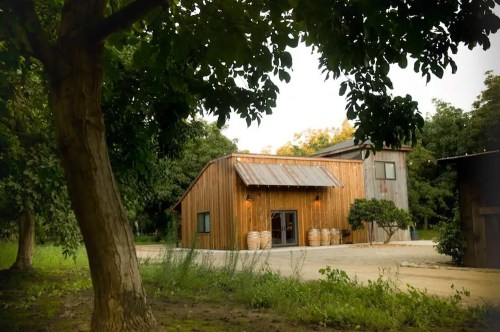 A rustic wooden building surrounded by trees, featuring a metal roof and barrels at the entrance.