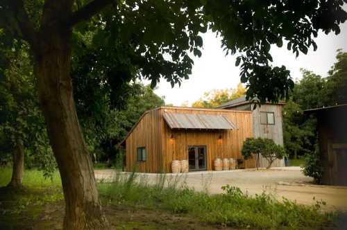 A rustic wooden building surrounded by trees, featuring a metal roof and barrels at the entrance.