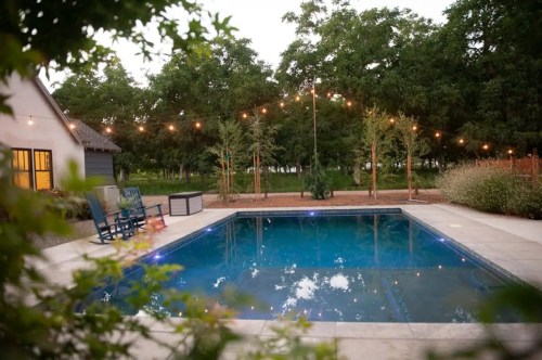 A serene backyard pool surrounded by trees and string lights, with a cozy seating area nearby.