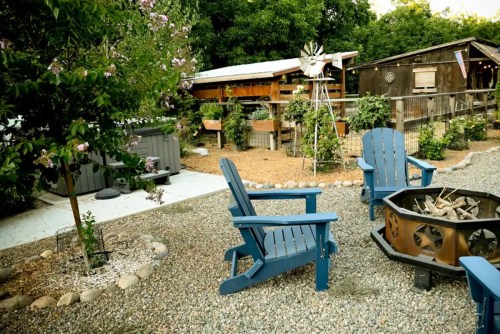 A cozy outdoor space with blue chairs, a fire pit, and greenery, featuring a rustic shed and a windmill in the background.