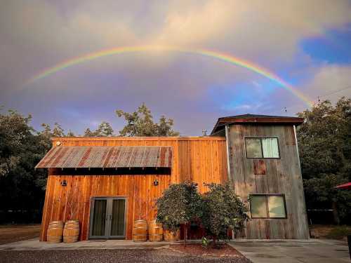 A rustic wooden house with a metal roof under a vibrant rainbow, surrounded by trees and barrels.