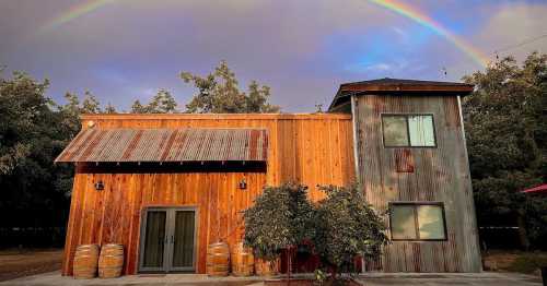 A rustic wooden building with a metal roof, surrounded by trees, under a vibrant rainbow in the sky.