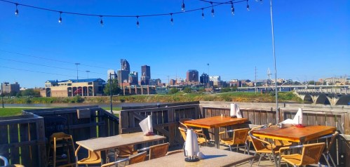 View of a city skyline from a rooftop patio with tables and string lights on a clear day.
