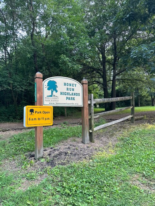 Sign for Honey Run Highlands Park, featuring park hours and a wooden fence in a green, wooded area.
