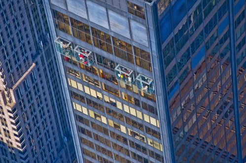 Workers in glass cleaning platforms on a tall skyscraper, with reflections of the city visible in the windows.