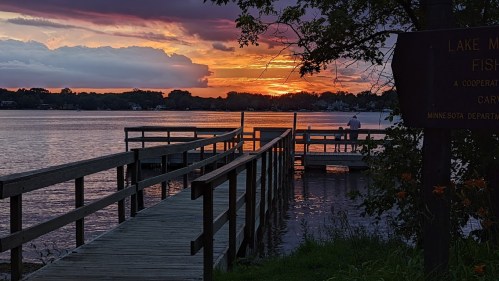 A serene sunset over a lake, with wooden docks extending into the water and a person sitting peacefully.