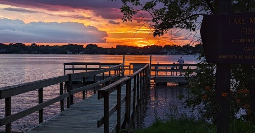 A serene sunset over a lake, with a wooden dock and a person fishing in the distance. Trees frame the scene.
