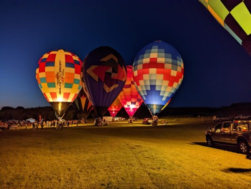 Colorful hot air balloons illuminated at dusk, with a crowd gathered on a grassy field.