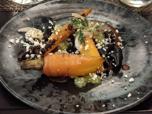 A gourmet dish featuring roasted carrots, herbs, and sauces on a dark plate, garnished with cheese and vegetables.