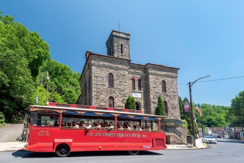A red trolley bus passes by a historic stone building on a sunny day, surrounded by greenery.