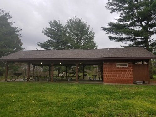 A covered picnic shelter with picnic tables, surrounded by trees and grass, under a cloudy sky.
