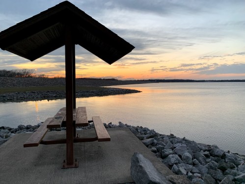 A picnic shelter by a calm lake at sunset, with rocks lining the shore and a colorful sky.