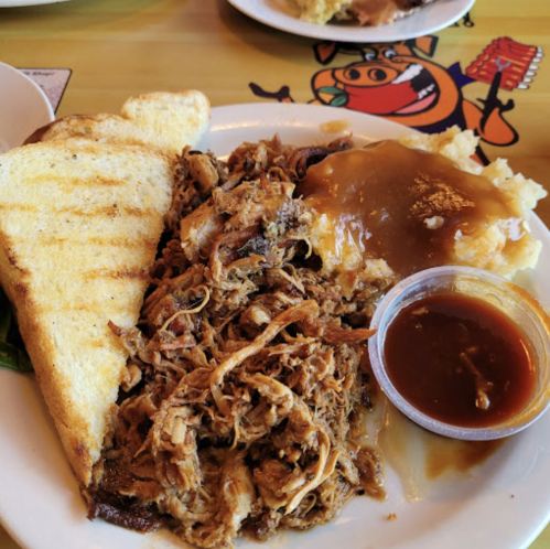 A plate of pulled pork with mashed potatoes and gravy, served with toasted bread and a side of barbecue sauce.