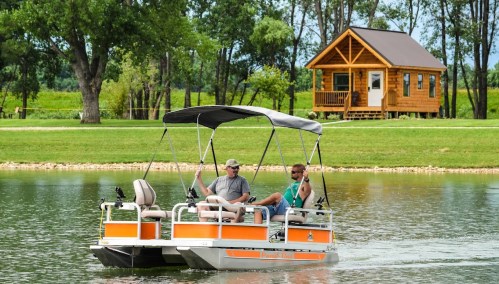 Two men relax on a small boat on a calm lake, with a cozy cabin visible on the shore in the background.