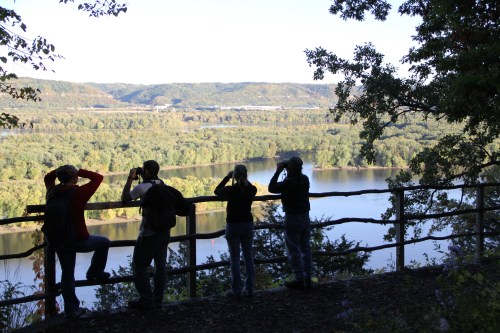 Four people silhouetted against a scenic river view, capturing the landscape with cameras from a wooded overlook.