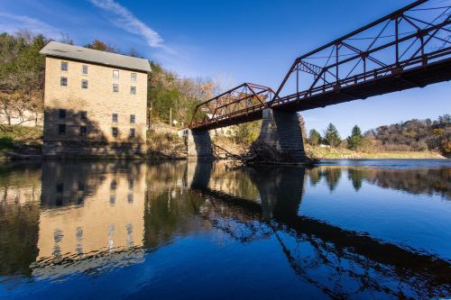 A stone building beside a river, with a metal bridge reflecting in the calm water under a clear blue sky.