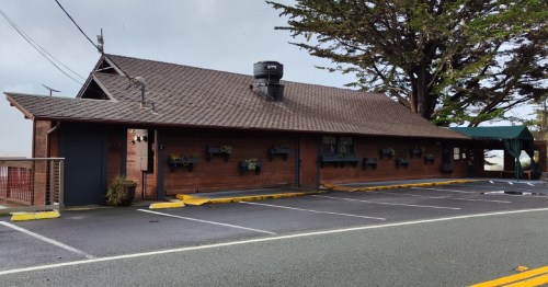 A wooden building with a sloped roof, featuring planters and a green awning, set beside a parking lot.