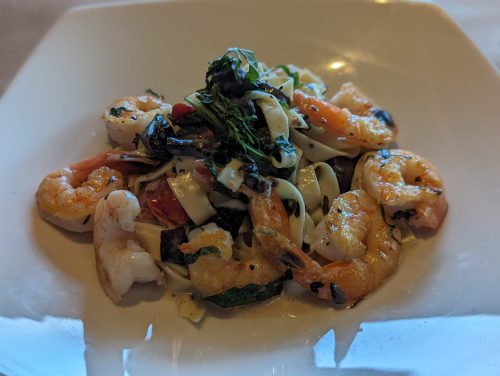 A plate of shrimp pasta with fettuccine, greens, and tomatoes, garnished and served in a white dish.