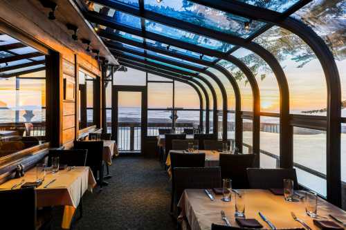 Cozy restaurant interior with glass walls, tables set for dining, and a stunning sunset view over the ocean.