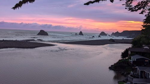 Sunset over a coastal landscape with rocky formations, gentle waves, and a calm river flowing into the ocean.