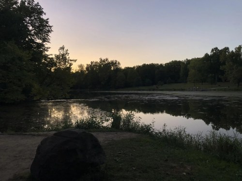 A serene lake at dusk, surrounded by trees, with a rock in the foreground and a calm reflection on the water.