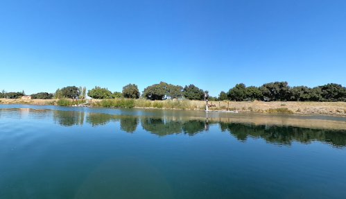 A calm river scene with clear blue skies, lush greenery, and reflections on the water's surface.