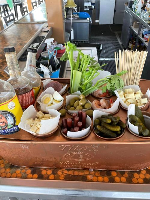 A variety of garnishes for cocktails, including olives, pickles, celery, cheese, and shrimp, displayed in a rustic container.