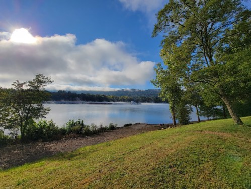 A serene lakeside view with trees, blue sky, and clouds reflecting on the water's surface.