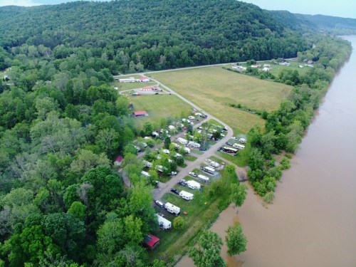 Aerial view of a riverside campground surrounded by lush greenery and hills, with RVs and tents arranged along a road.