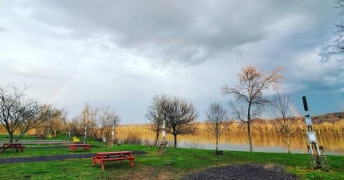 A serene landscape featuring a river, bare trees, picnic tables, and a rainbow in a cloudy sky.